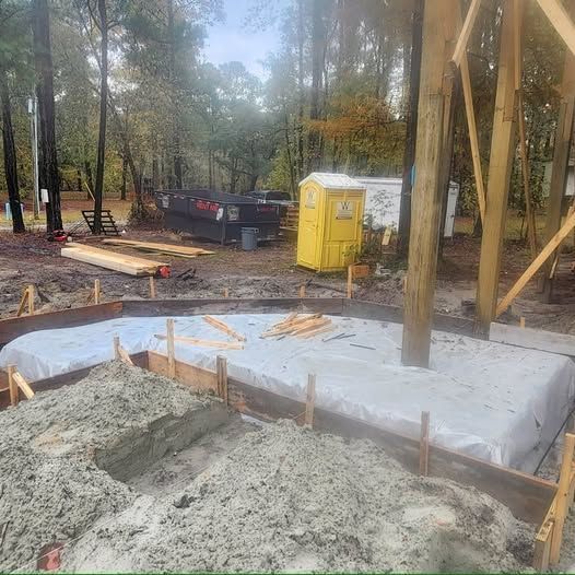 Construction site with wooden frame and concrete base, trees in background, yellow portable toilet.