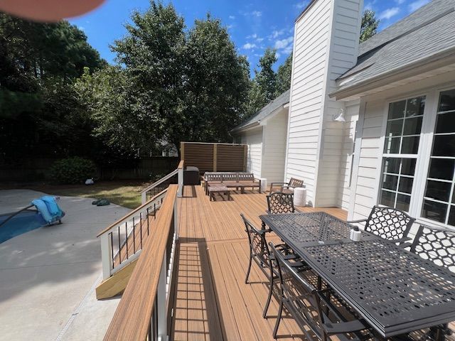 Wooden deck with outdoor furniture next to a white house with a chimney and surrounding trees.