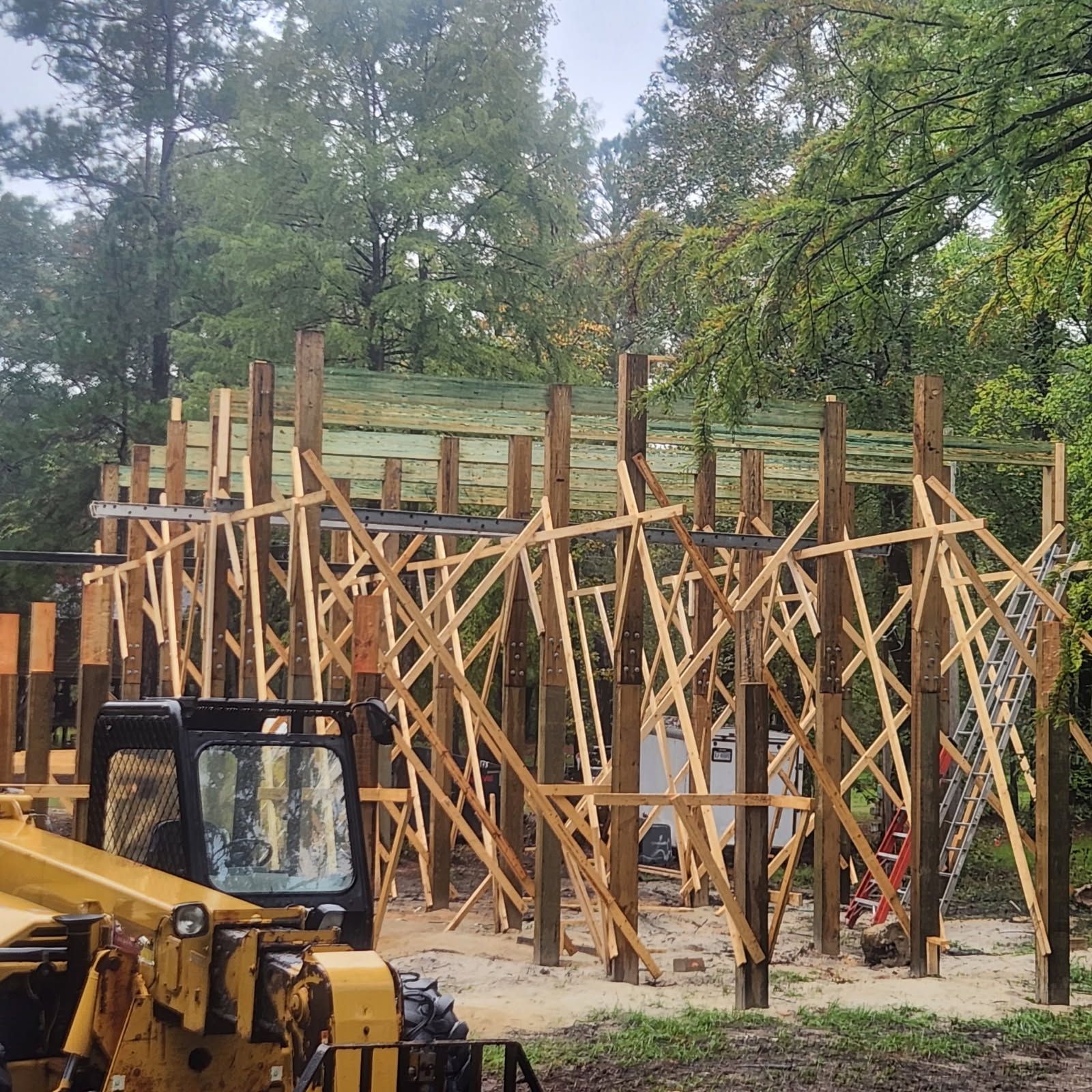 Construction of a wooden building frame. A bulldozer is parked nearby. Trees are in the background.