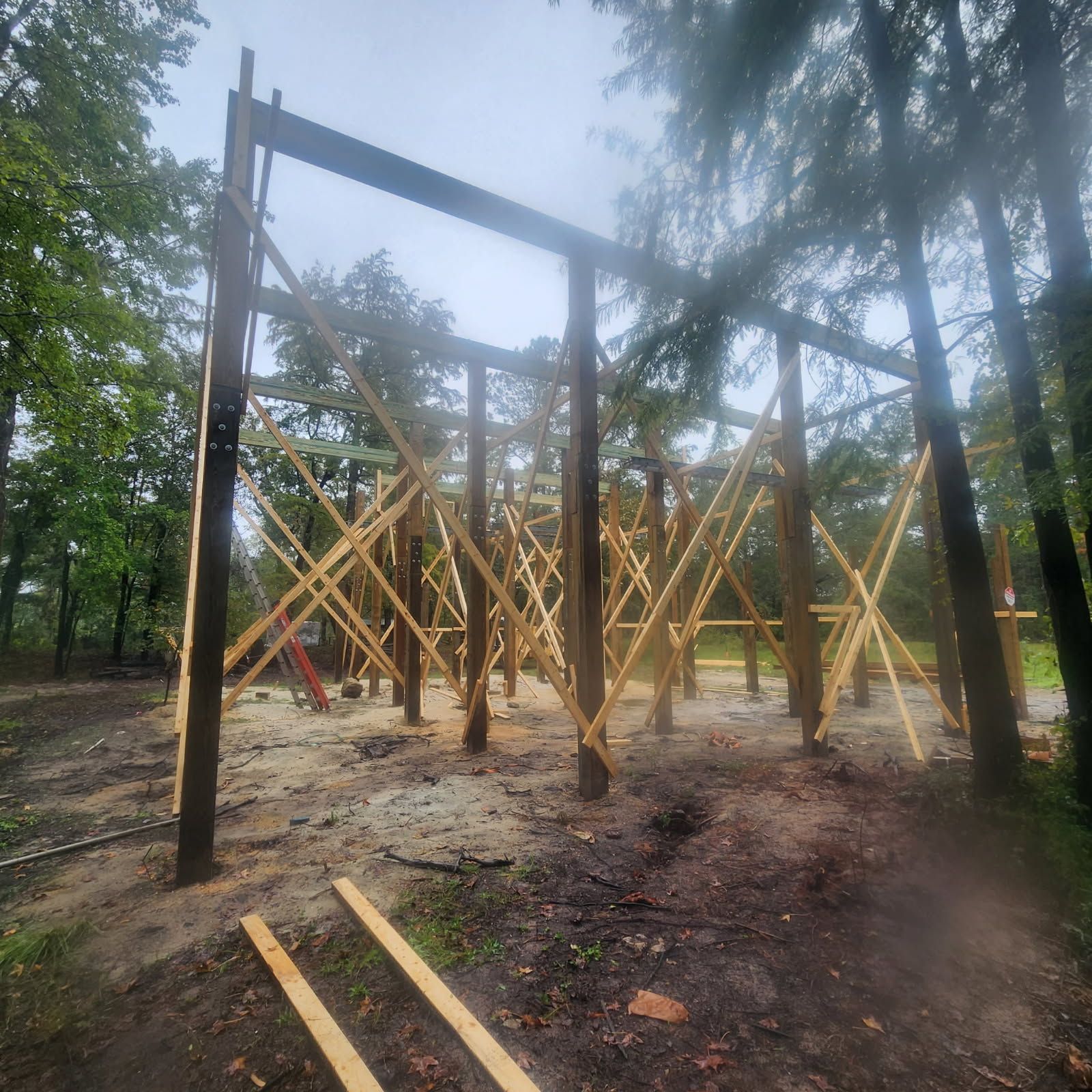 Wooden frame structure being built in a wooded area, with cross-bracing and a partially built roof.