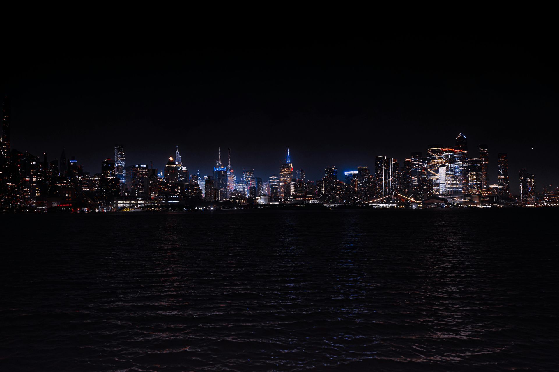 The illuminated skyline of a city at night, reflecting over a calm body of water.