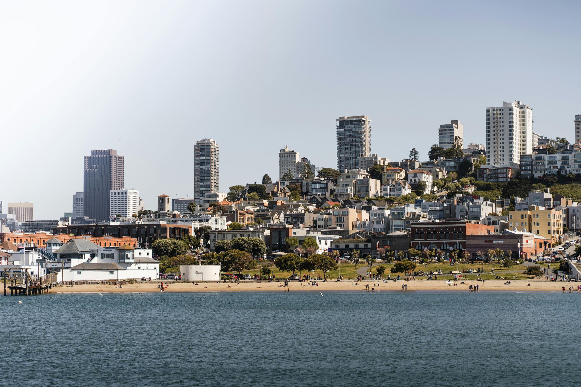 A sunny, wide view of a city skyline rising above a sandy beach and calm water.