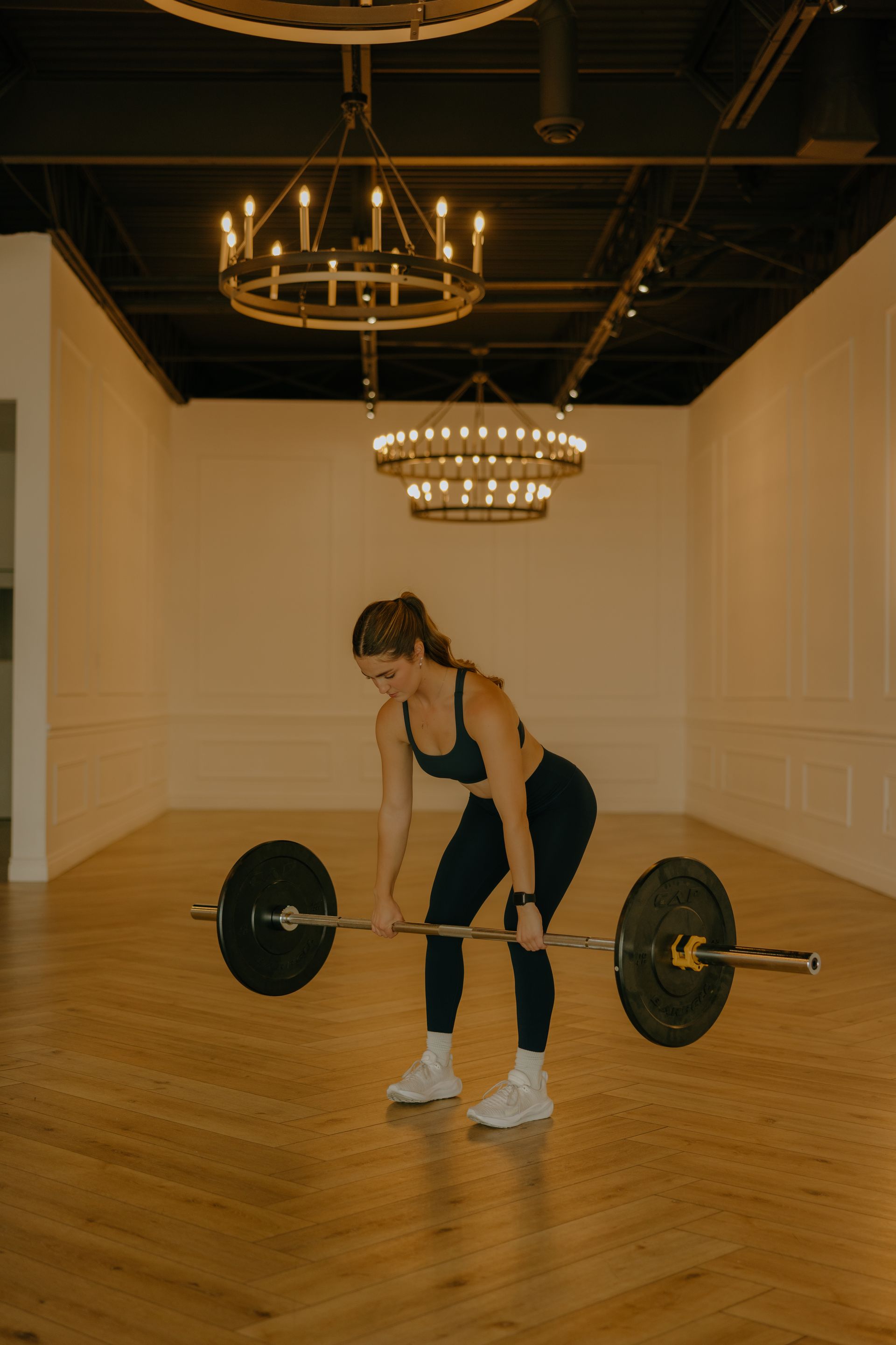 Woman in gym gear deadlifting a barbell in a bright, open room with wooden floors and decorative chandeliers.
