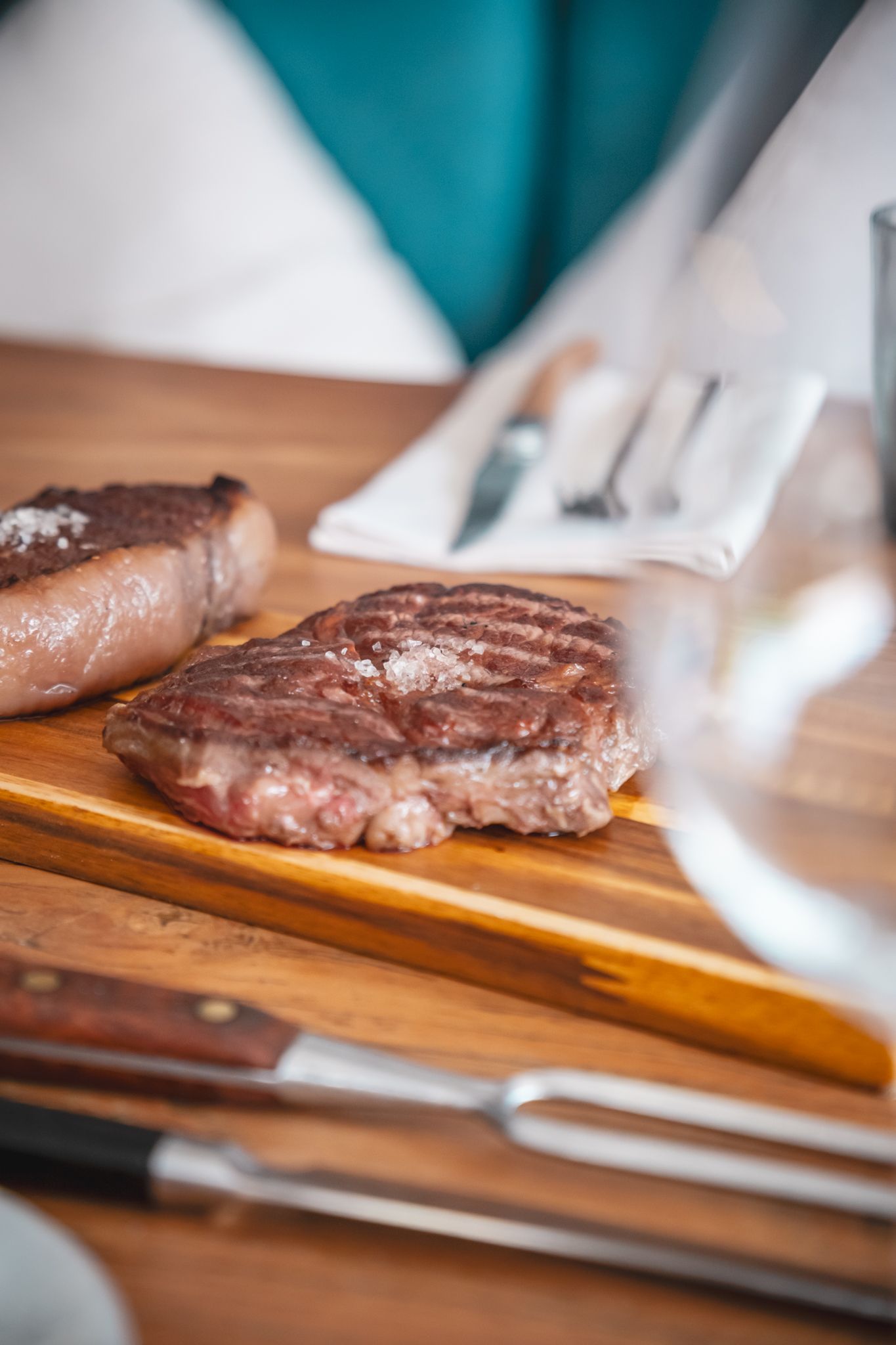 Steak cooking on a wooden cutting board with utensils beside it in a kitchen setting