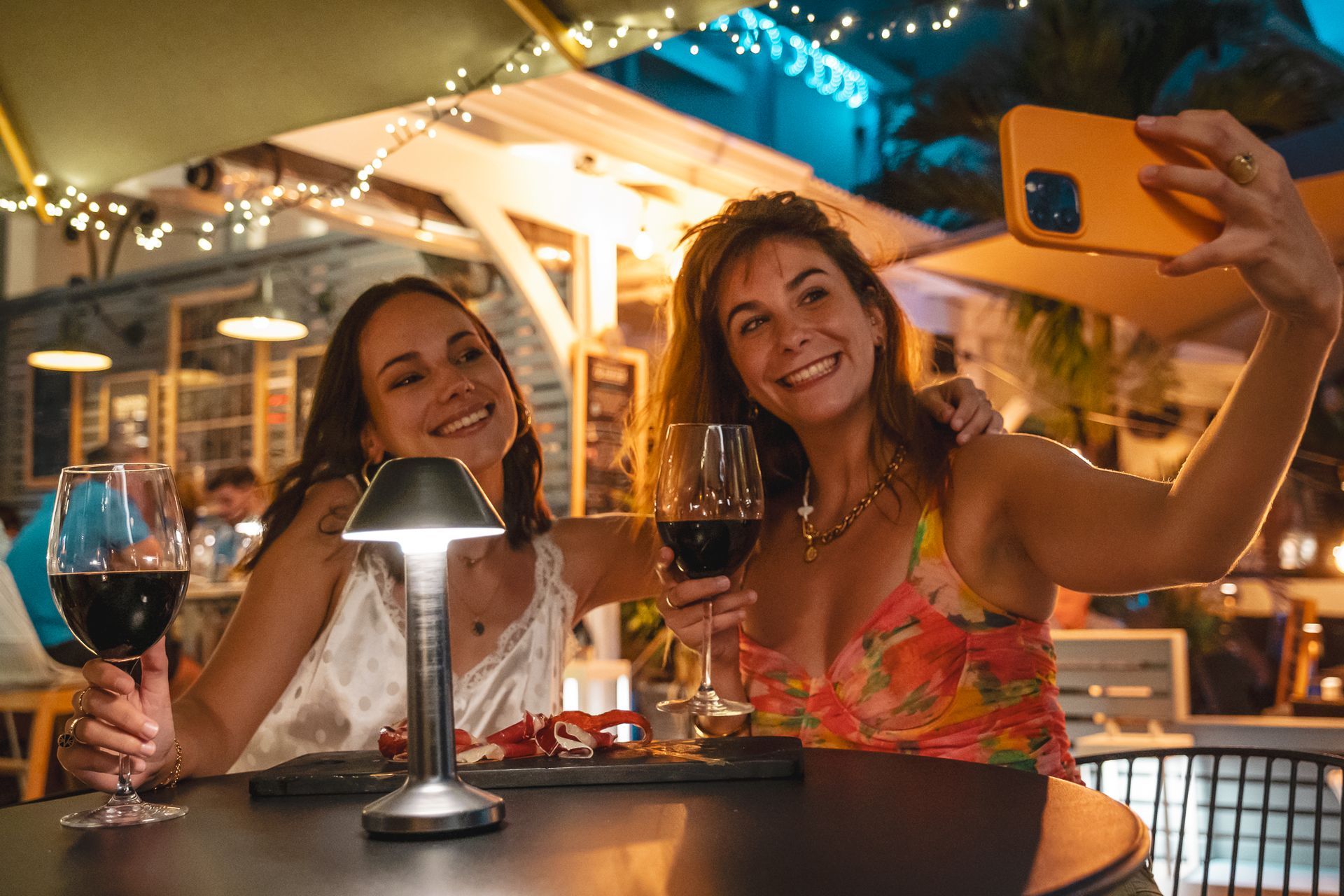 Two friends smiling at a restaurant table, taking a selfie with wine glasses and a lit lamp at night