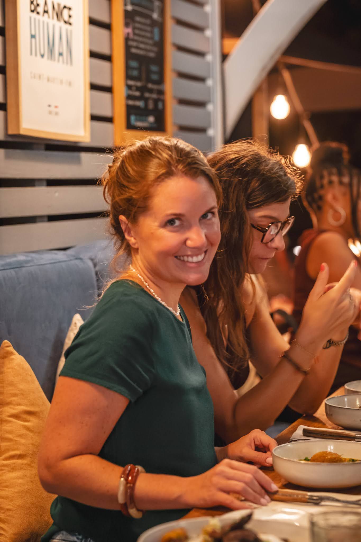 Woman smiling at a restaurant table with plates and drinks, seated beside another diner in a cozy setting