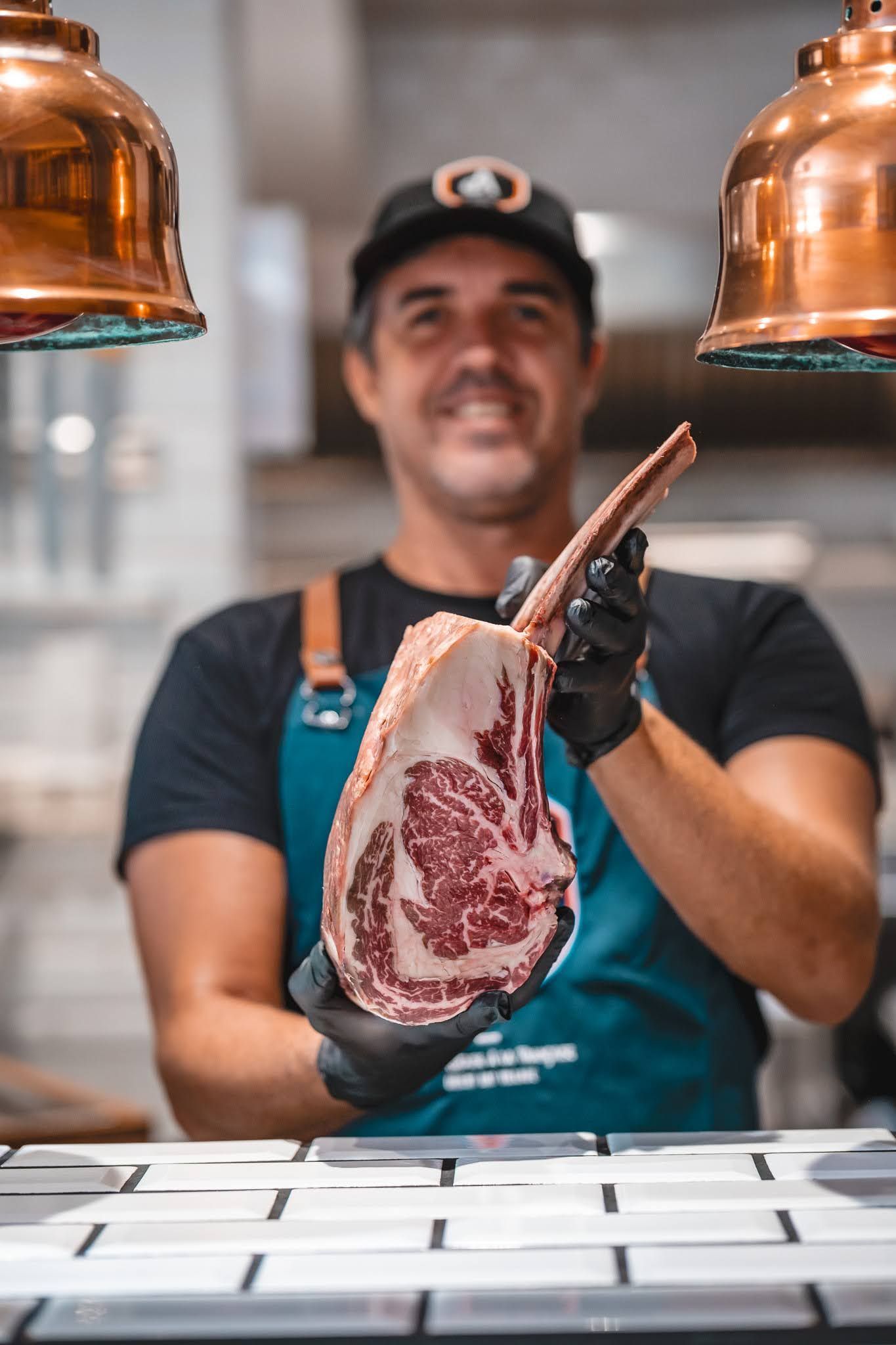 Butcher holding a hanging cut of beef in a market stall under copper heat lamps