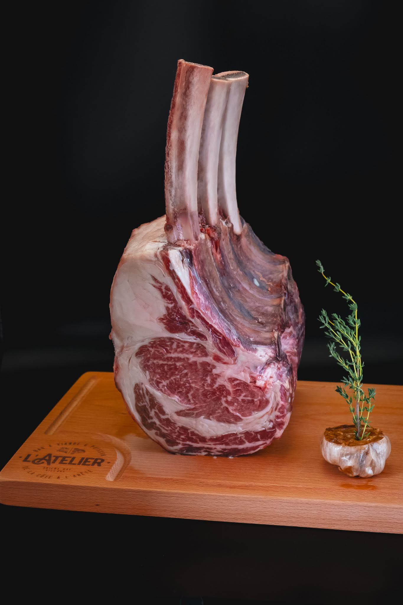 A raw rib roast on a wooden board beside a small herb sprig against a black background