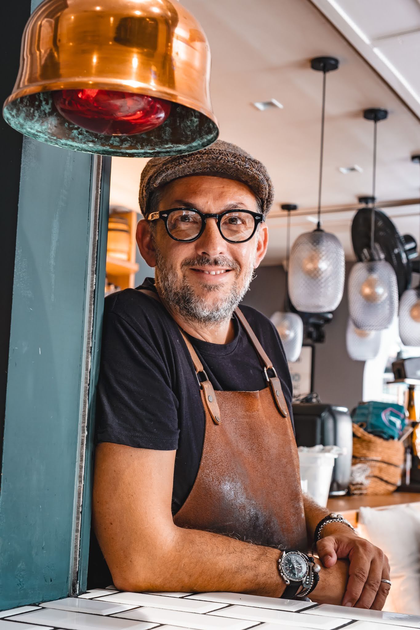 Smiling café worker in glasses and apron standing behind a counter under hanging pendant lights
