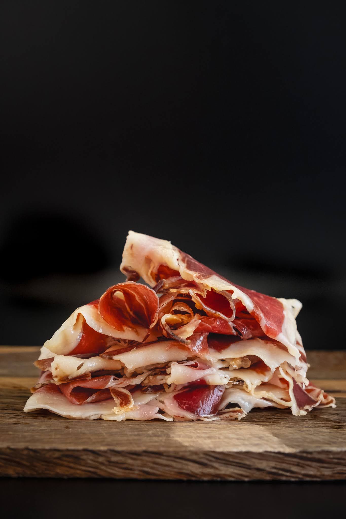 Stack of thinly sliced cured meat on a wooden board against a dark background