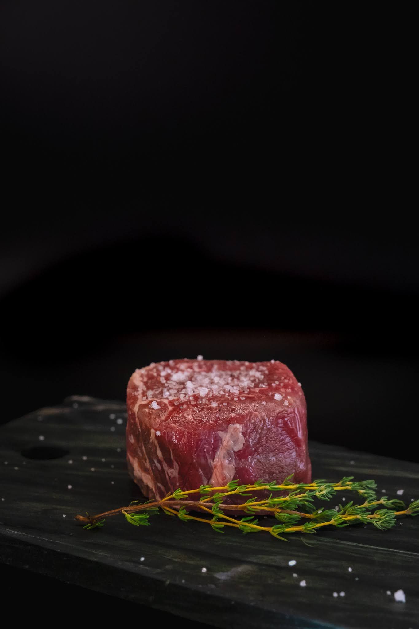 Raw steak on a dark plate with green garnish, shot against a black background