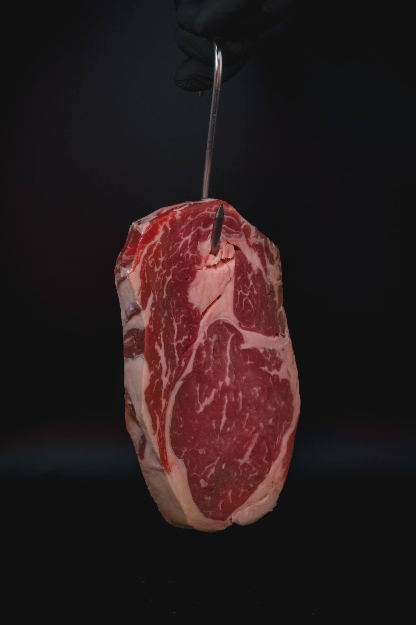 Raw marbled steak hanging from a metal hook against a dark background