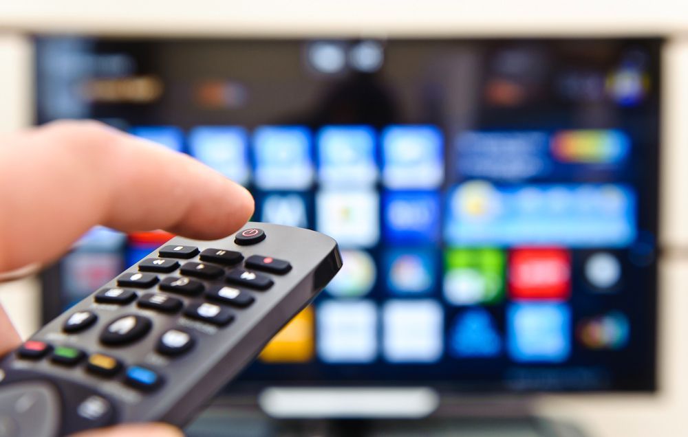 Hand holding TV remote, pointing it at a flat-screen TV displaying a colourful app menu - Powered By JBES In Sandgate, NSW