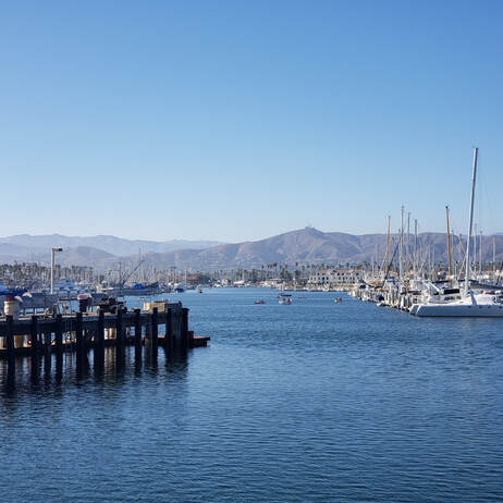 Boats docked in a harbor under a clear blue sky, with mountains in the background.