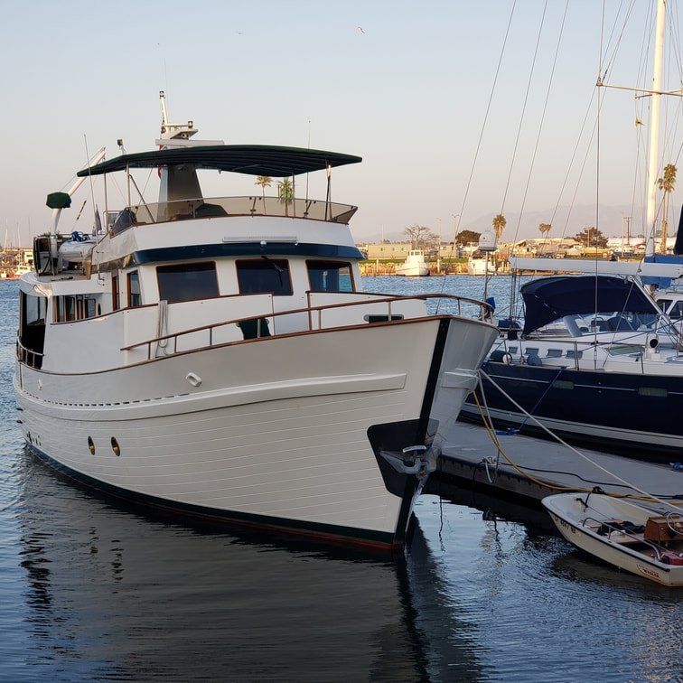 White motor yacht docked in harbor, next to smaller sailboats. Blue water, sunny day.