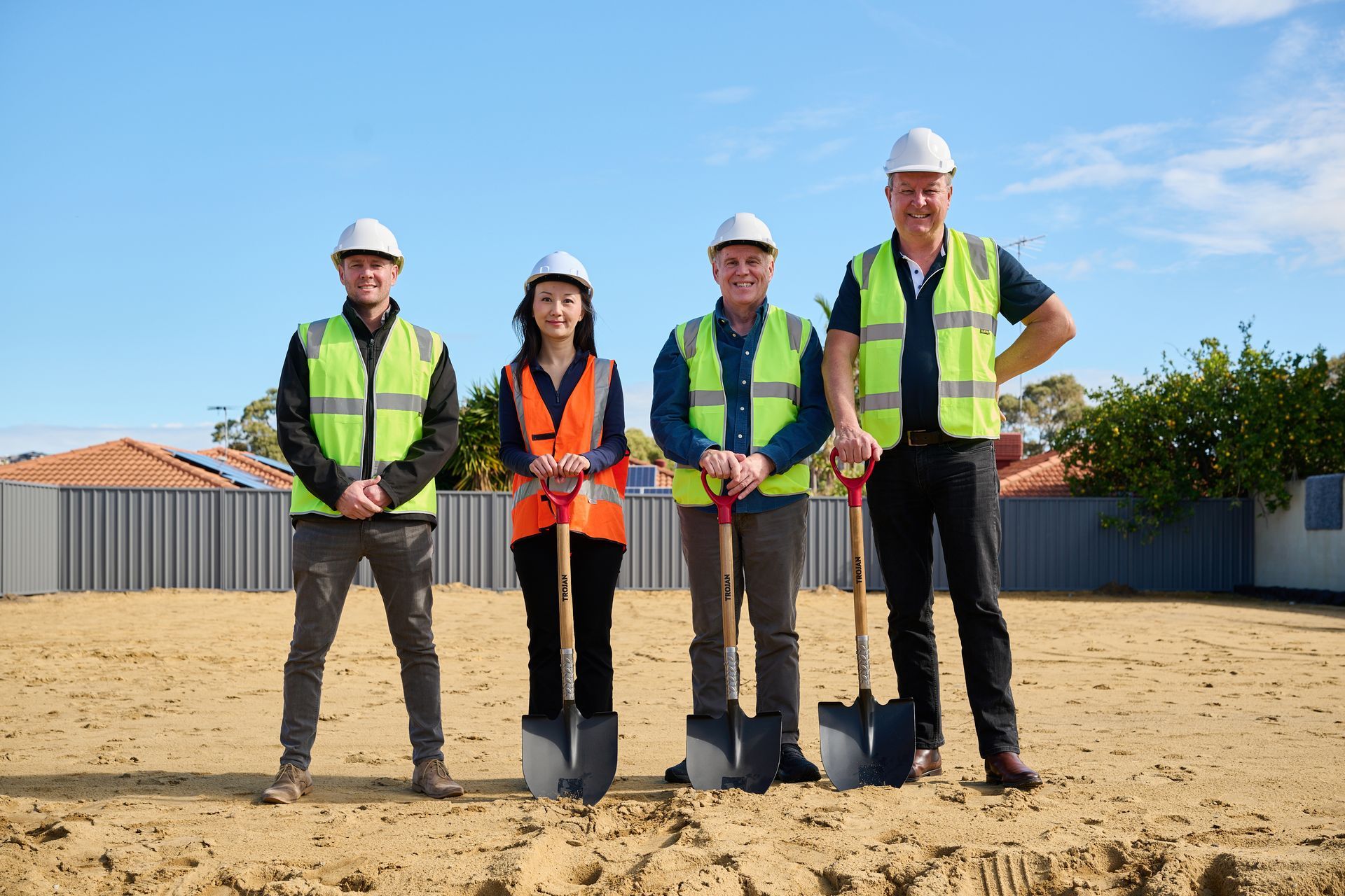 Four construction workers in safety vests and hard hats stand on a sandy lot with shovels.