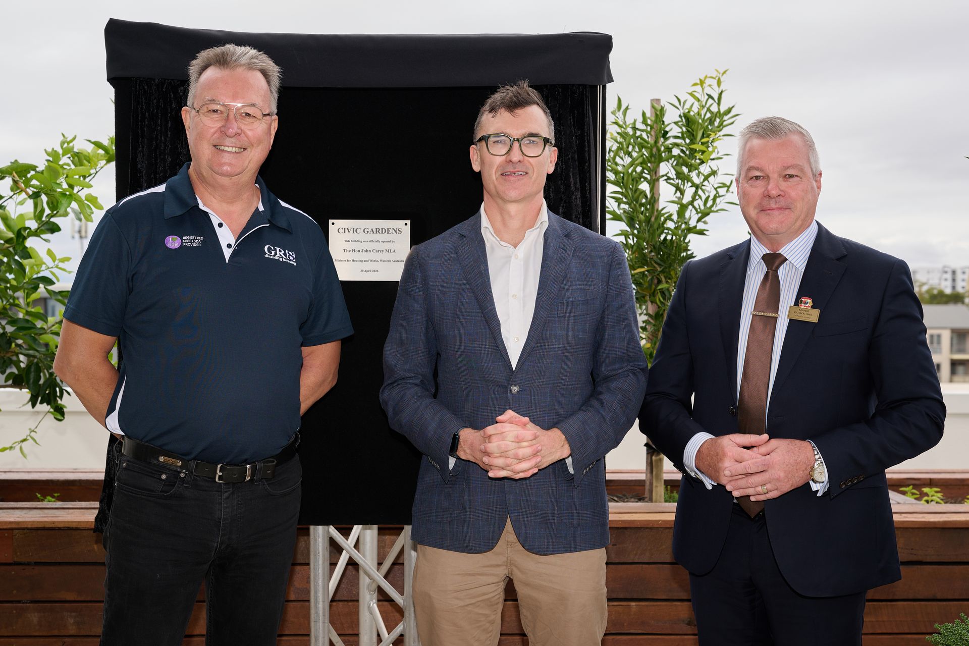 Three men in suits posing outdoors in front of a black display board and plants