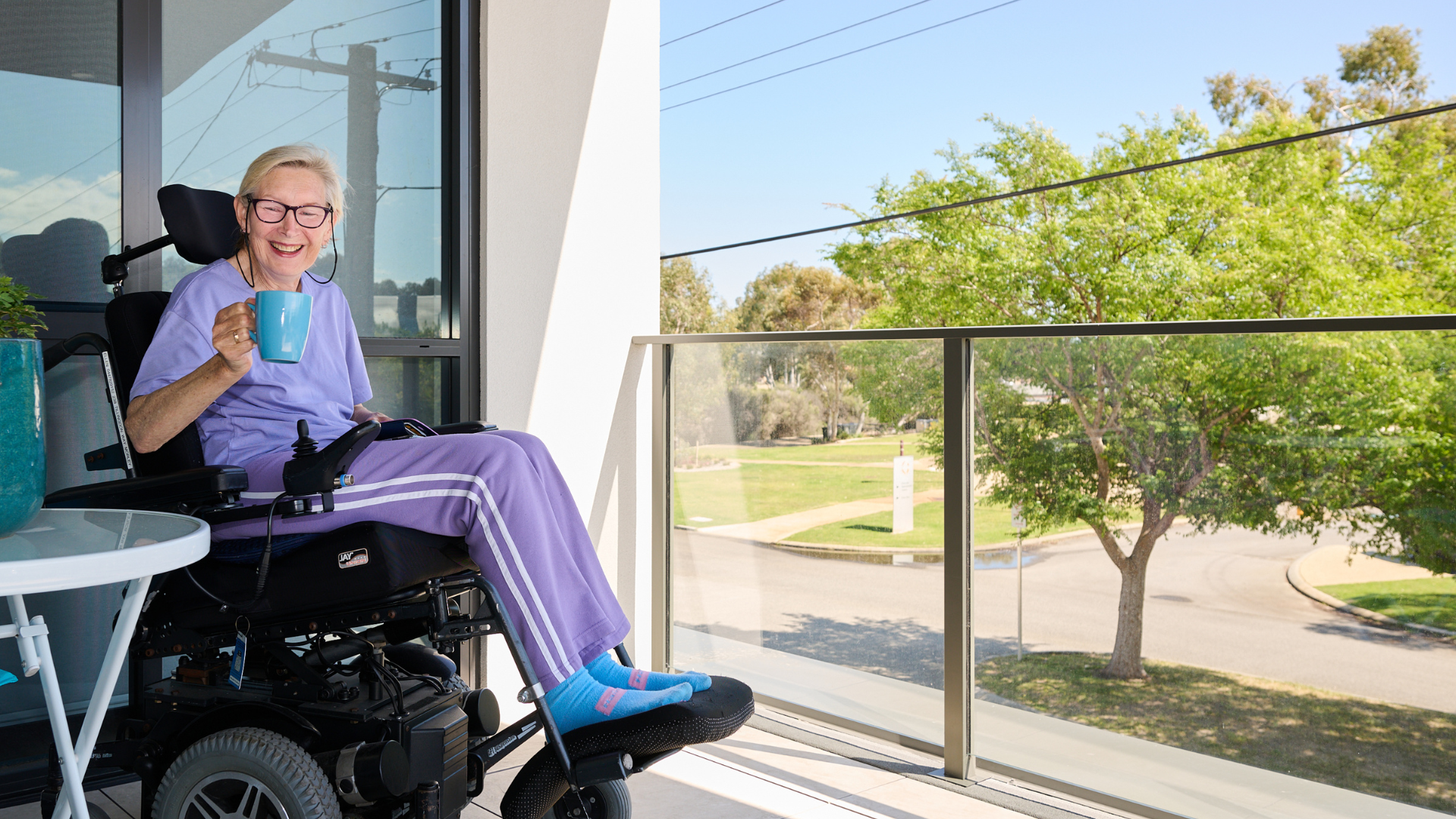 Person in wheelchair on a balcony, smiling and holding a drink, with trees and a street view beyond the railing