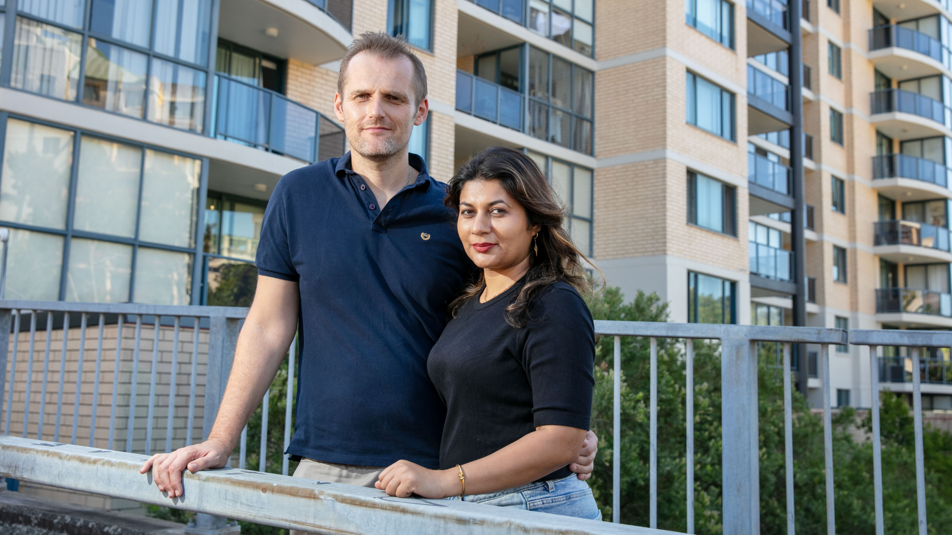 Two people leaning on a railing in front of modern apartment buildings.