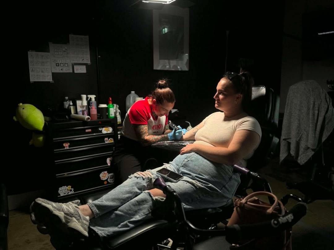 Tattoo artist working on a seated client in a dim studio, with tools and supplies on a nearby cart