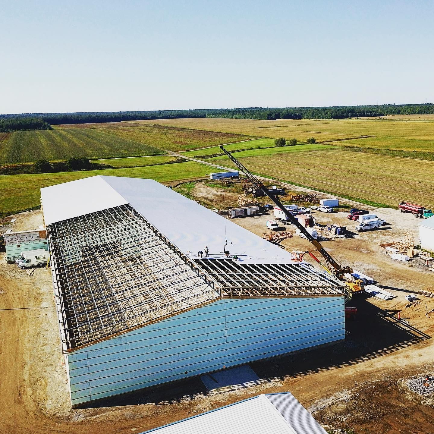 Large industrial building under construction, exterior view with blue siding, metal roof, and surrounding fields.