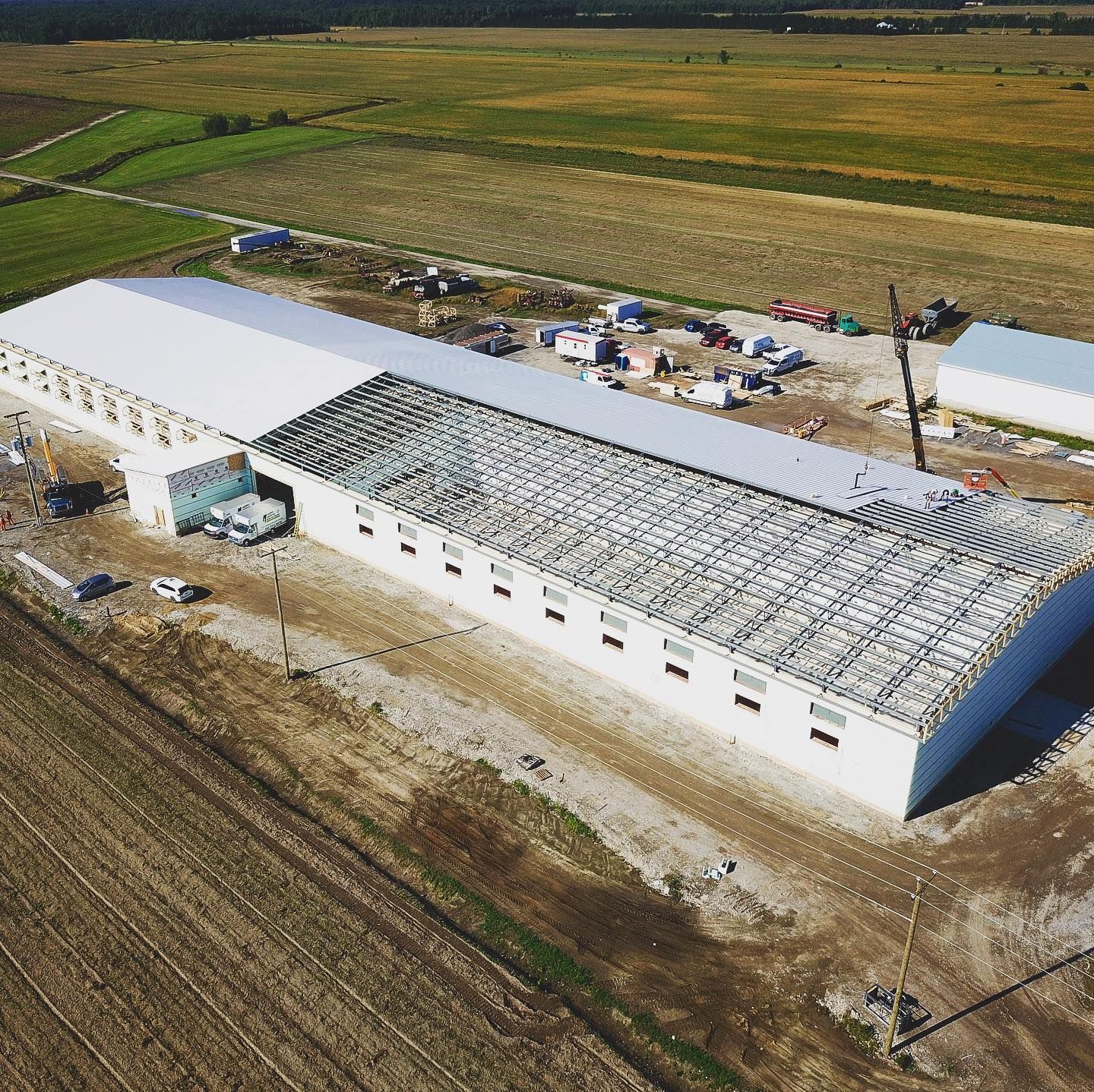 Aerial view of a long, white industrial building with a curved roof in a field, construction ongoing.