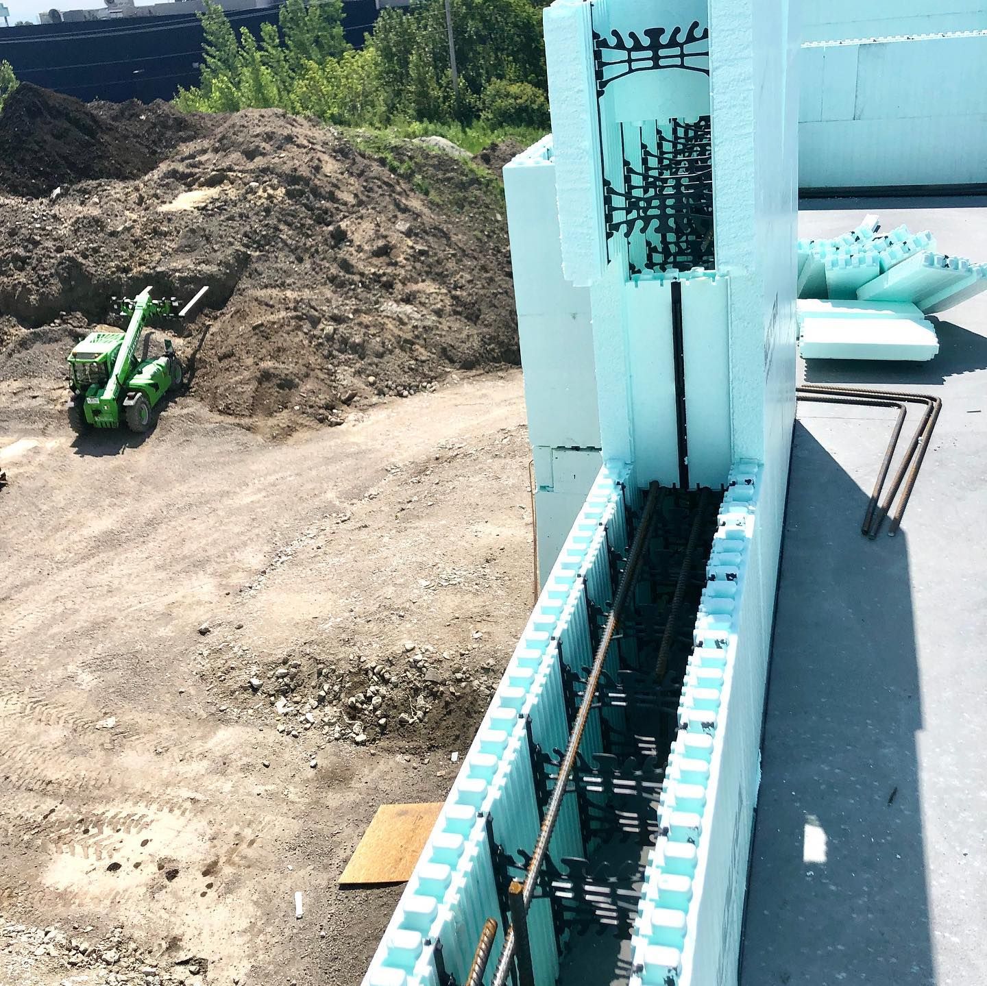 Construction site with ICF walls, rebar, and earth-moving equipment. Blue foam blocks, tan dirt, and green machinery.