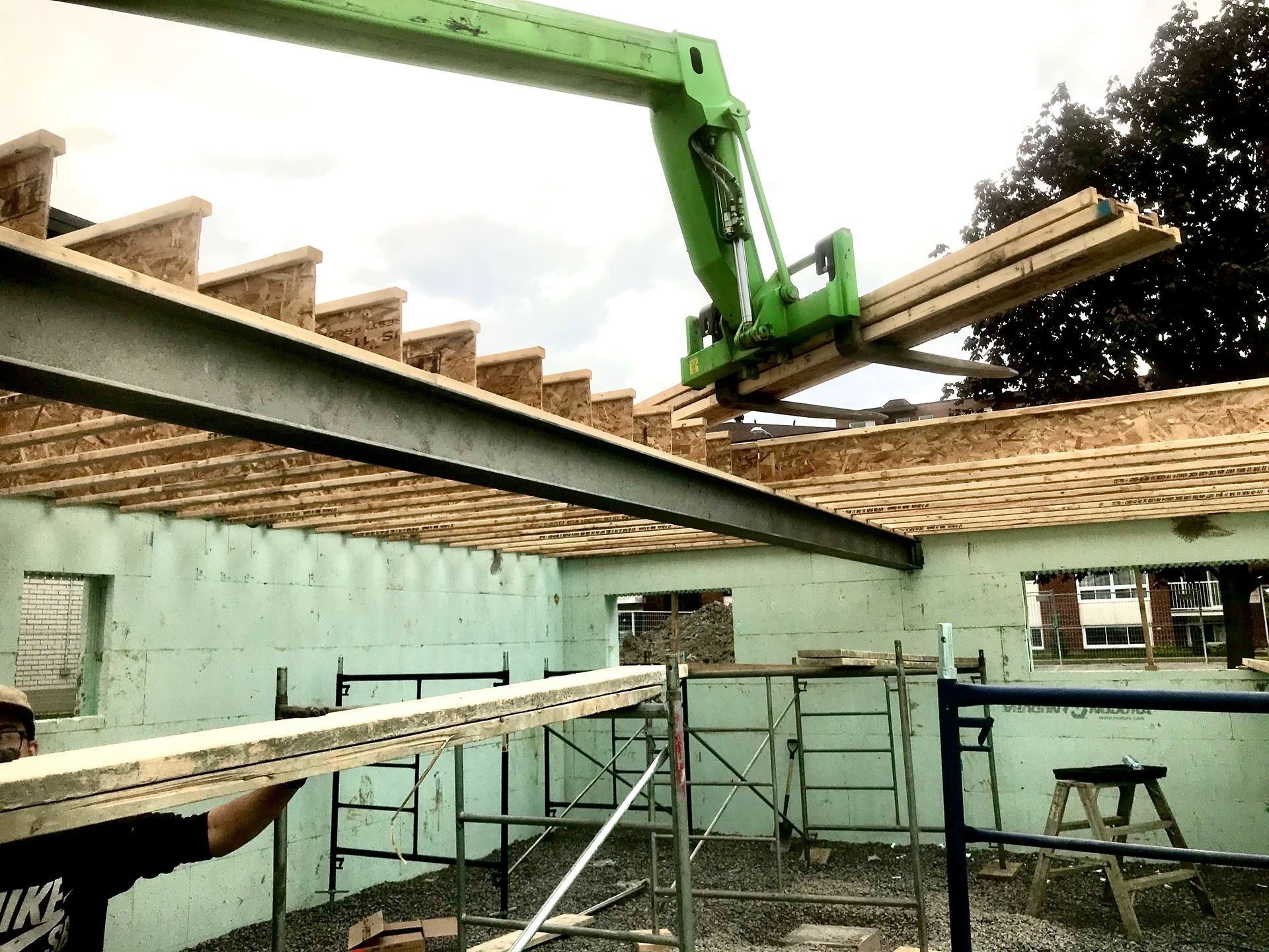 Construction site: Green lift arm placing wood beams onto roof framework. Man holding wood.