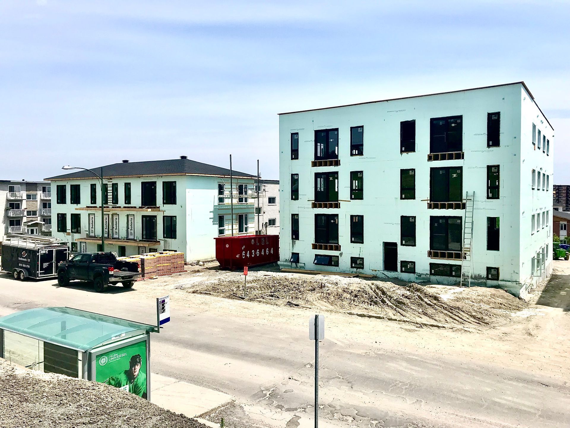 Two light-colored, modern buildings under construction on a dirt lot. The sky is blue.