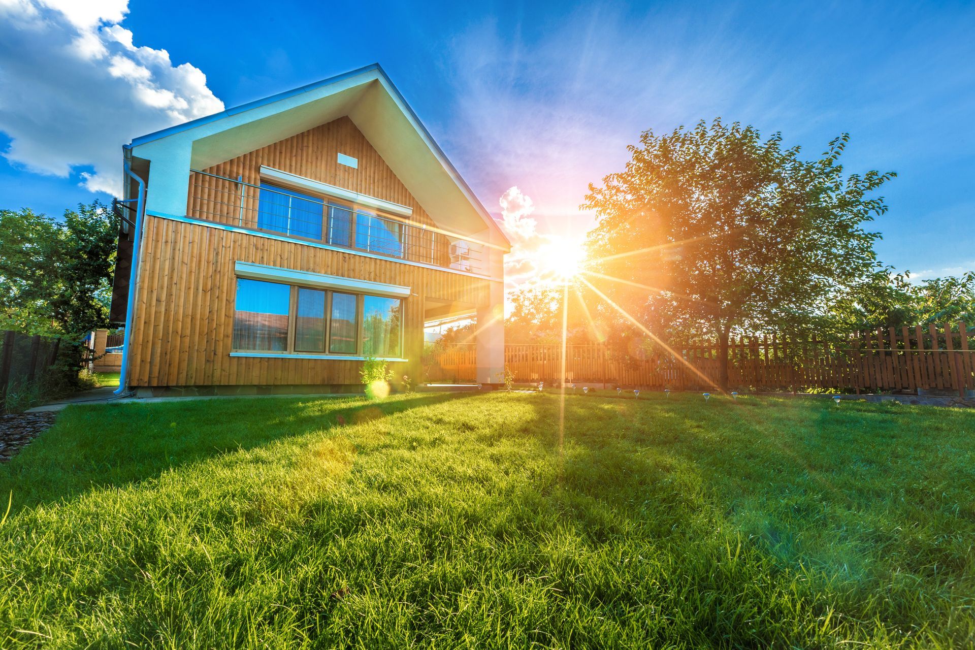 Moderna casa in legno con prato verde lussureggiante, luce solare intensa e cielo azzurro.
