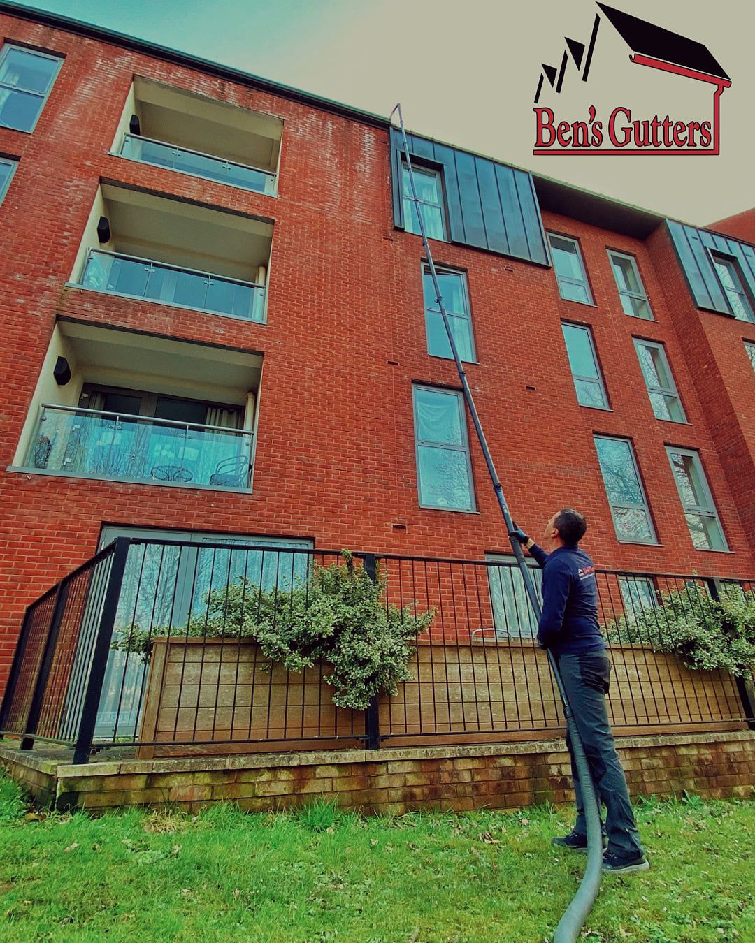 Man cleaning windows on a red brick building using a water-fed pole. Green grass and shrubs in the foreground.