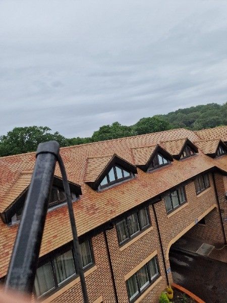 Brown tiled roof with dormer windows on a brick building, set against a cloudy sky and trees.