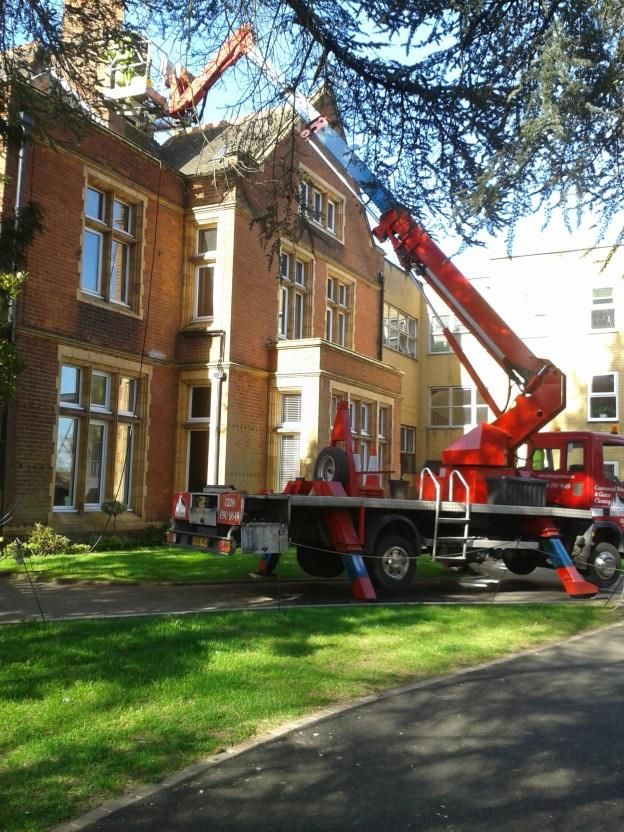 Red crane truck working on the roof of a brick building. Trees and grass in the foreground.