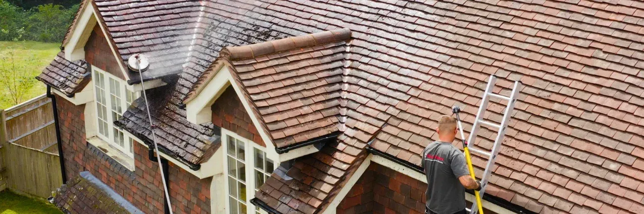 Man on a ladder cleaning a roof of a brick house. The roof is made of brown tiles.