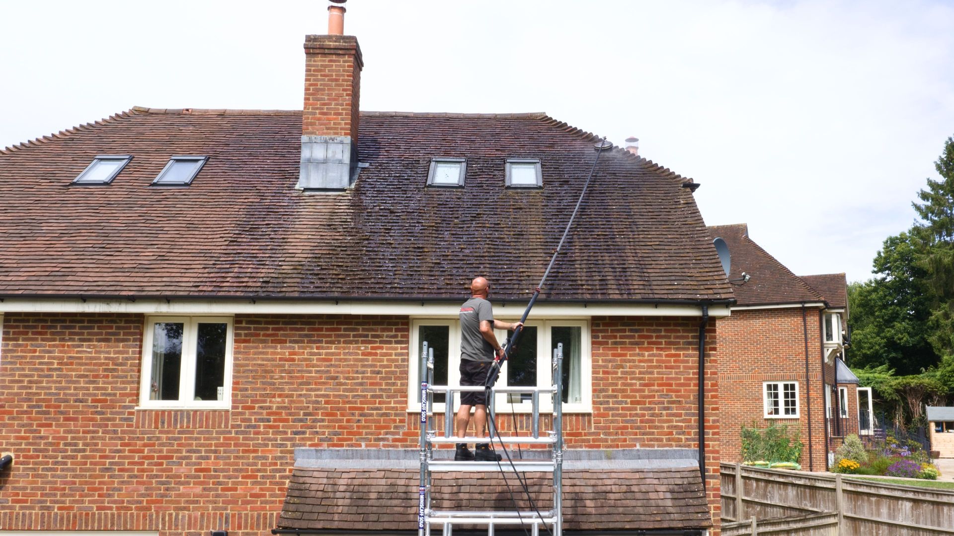 Man on ladder power washing a brick house roof with moss.