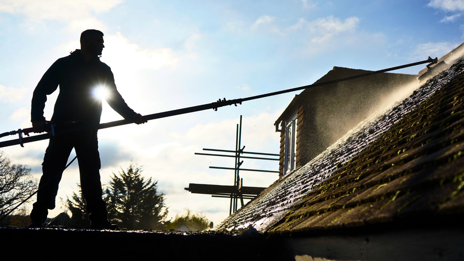 Person washes a roof with a long hose, backlit by the sun.