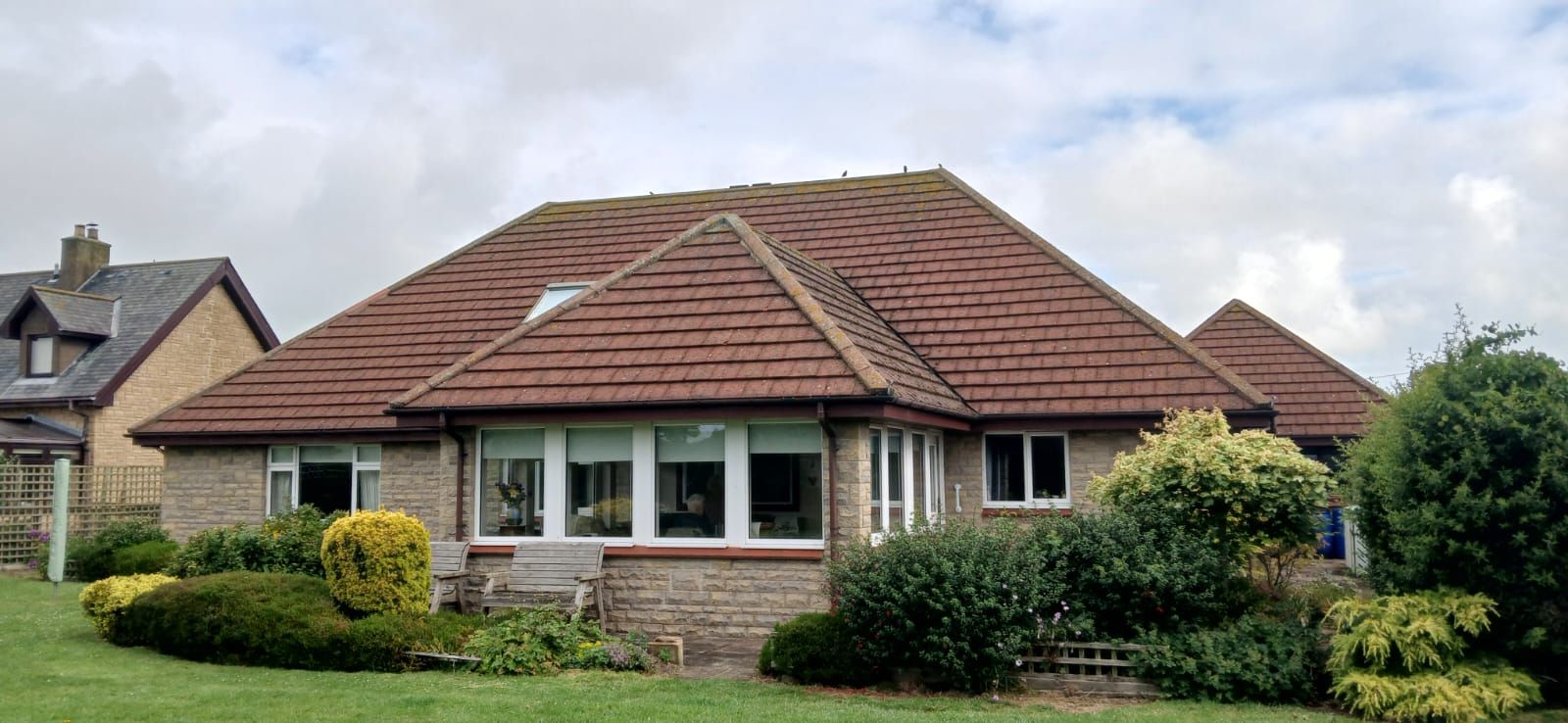 A single-story brick house with a red tiled roof, surrounded by green shrubs and grass, under a cloudy sky.