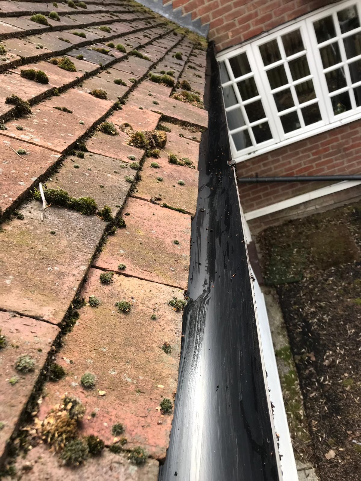 Red tile roof with moss, and black gutter next to a brick wall with a white window.