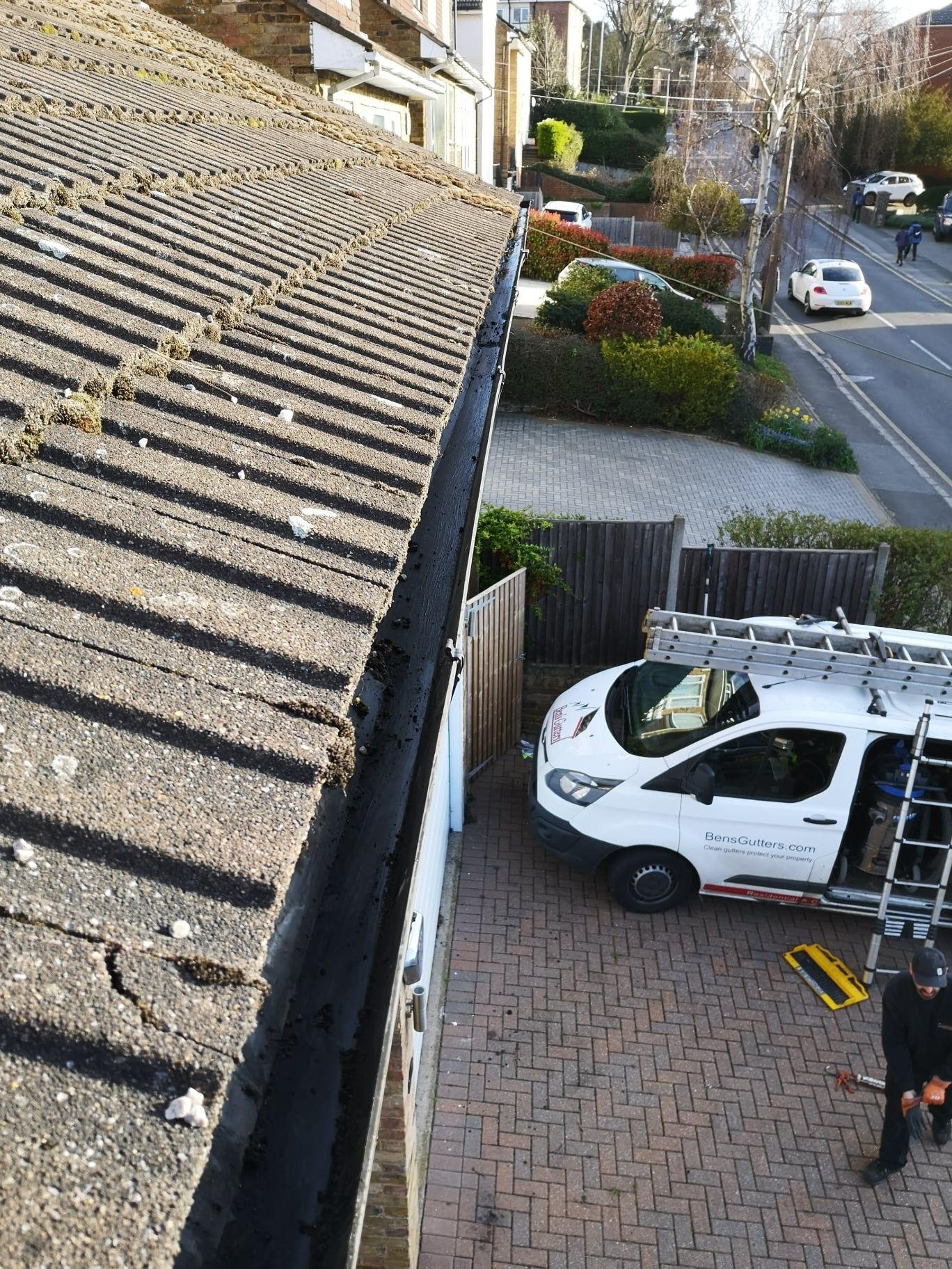 Gutter cleaning service: white van with ladder in driveway.  Workers near roof, next to a street.