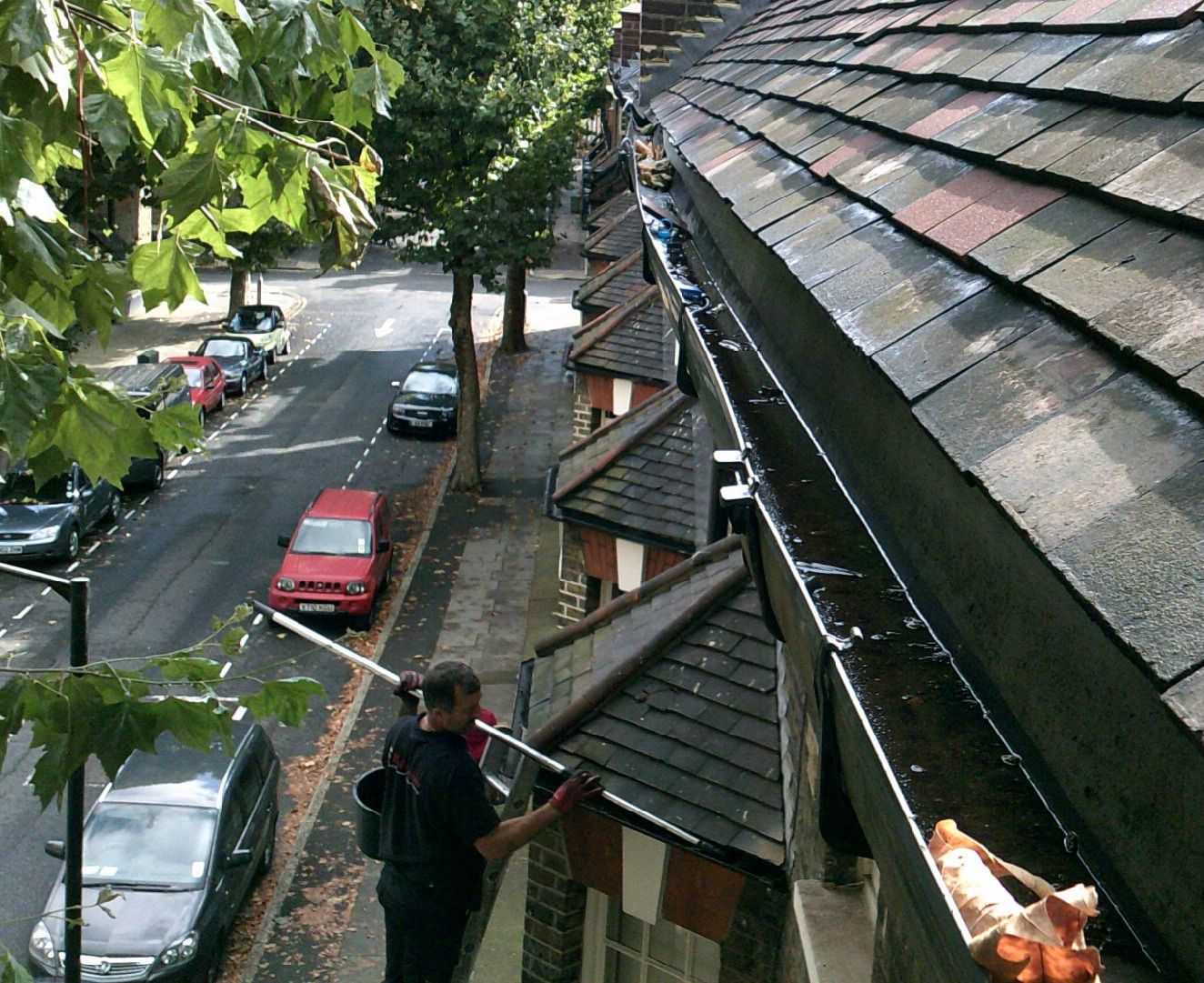 Man cleaning a rooftop gutter on a building overlooking a street with parked cars.