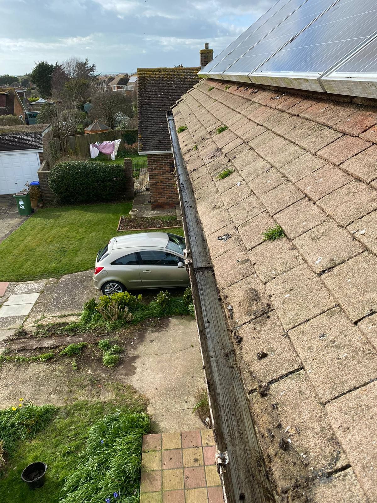 View of a house rooftop with gutters, weeds, and solar panels. A car is parked on the street below.