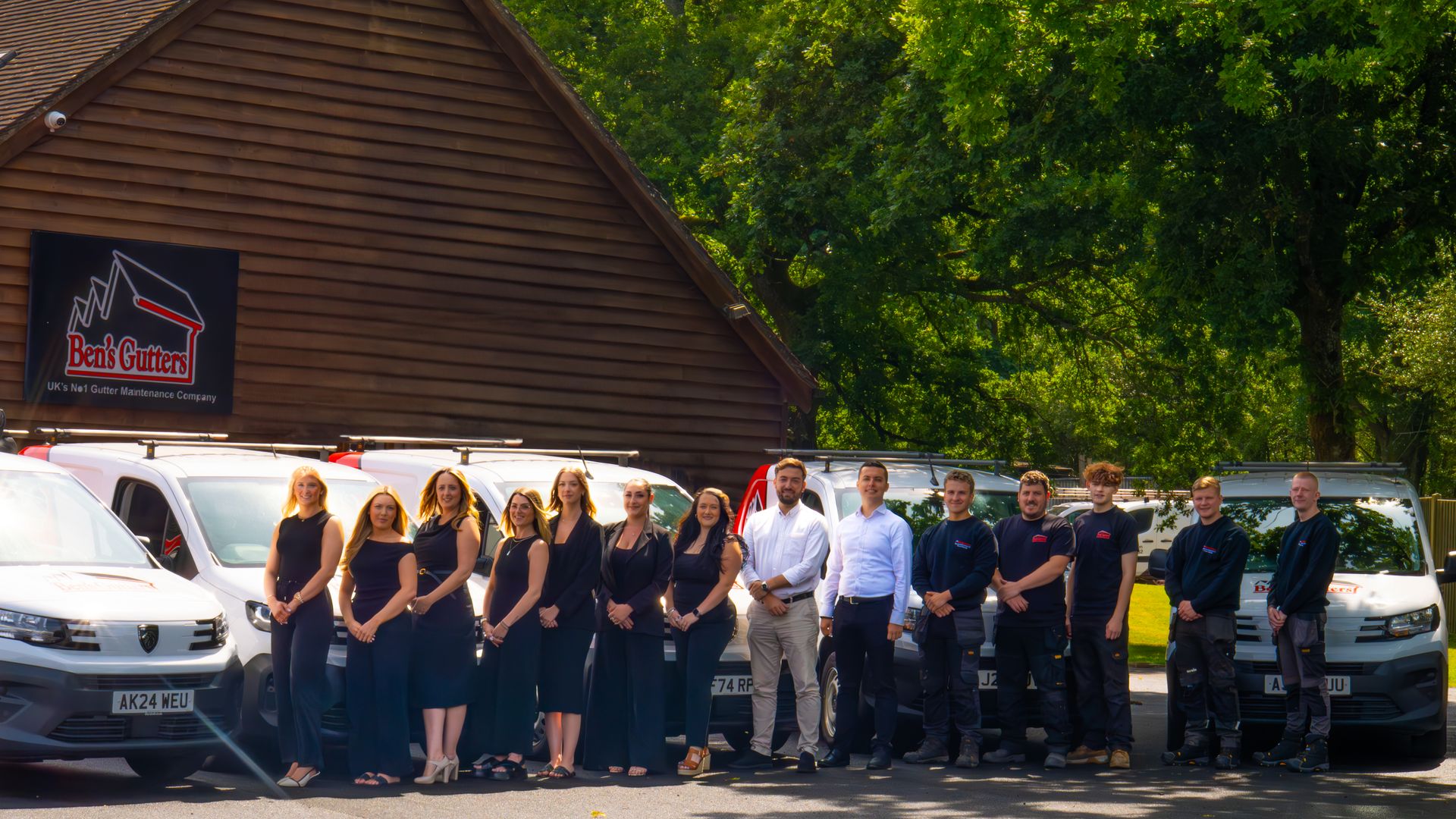 Group of people standing in front of vans and a building with a logo; sunny outdoor setting.