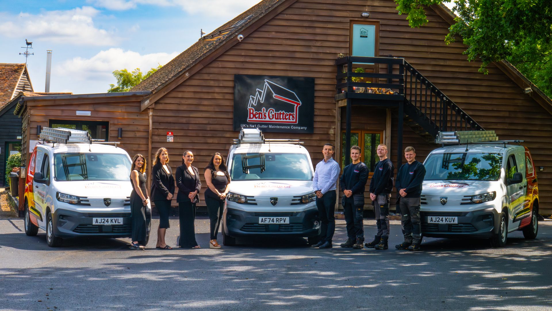 Group of people standing with three vans in front of a wooden building.