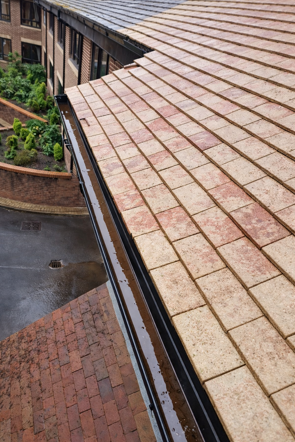 Red and tan tiled roof with black gutters, viewed from above, with building and brick wall in background.