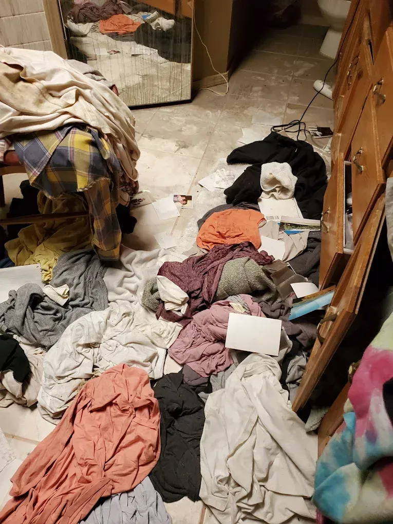 Clothes scattered across a bedroom floor beside a dresser and bed, suggesting a messy room.