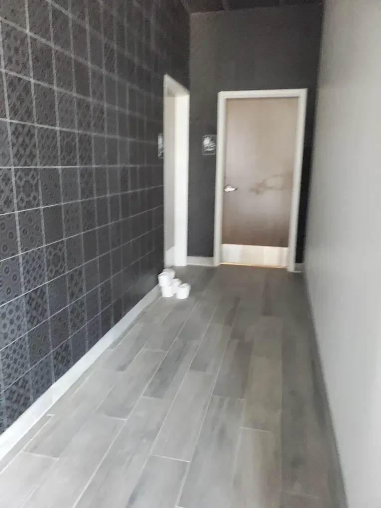 Narrow hallway with gray tile floor, dark tiled left wall, white right wall, and a closed door at the end