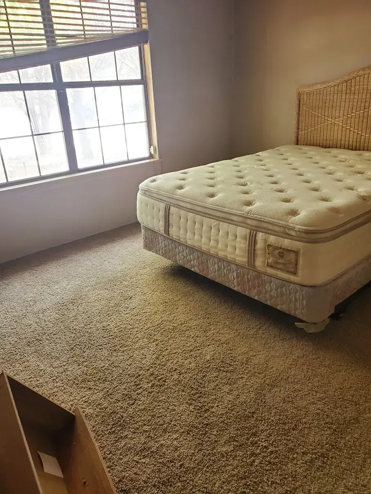 Sparse bedroom with beige carpet, window blinds, and an unmade white mattress on a box spring