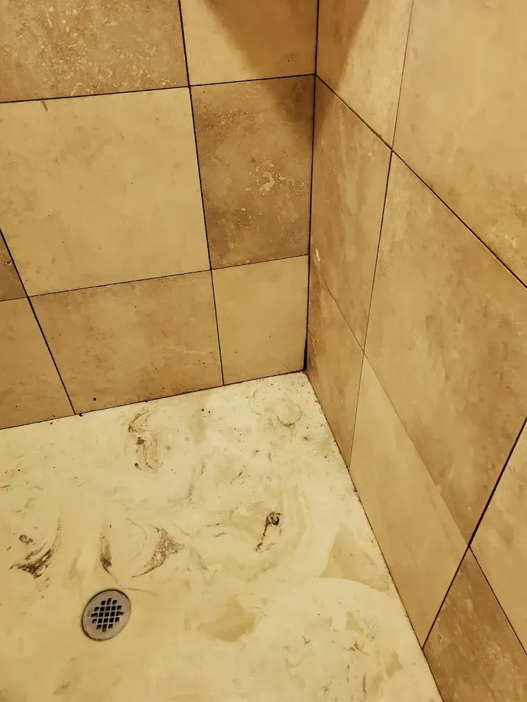 Tiled shower corner with dirty beige walls and floor, showing a drain and stained grout