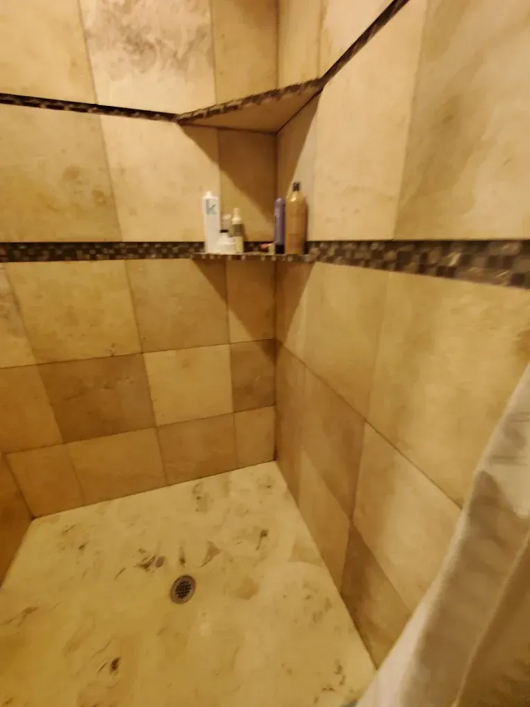 Beige tiled shower corner with a drain, shelf, and toiletries on the wall.