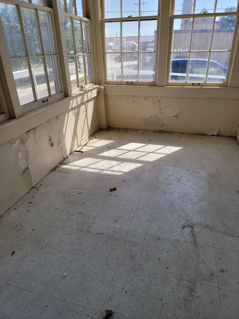 Empty sunlit room with large windows, cracked white walls, and worn floor casting window shadows.